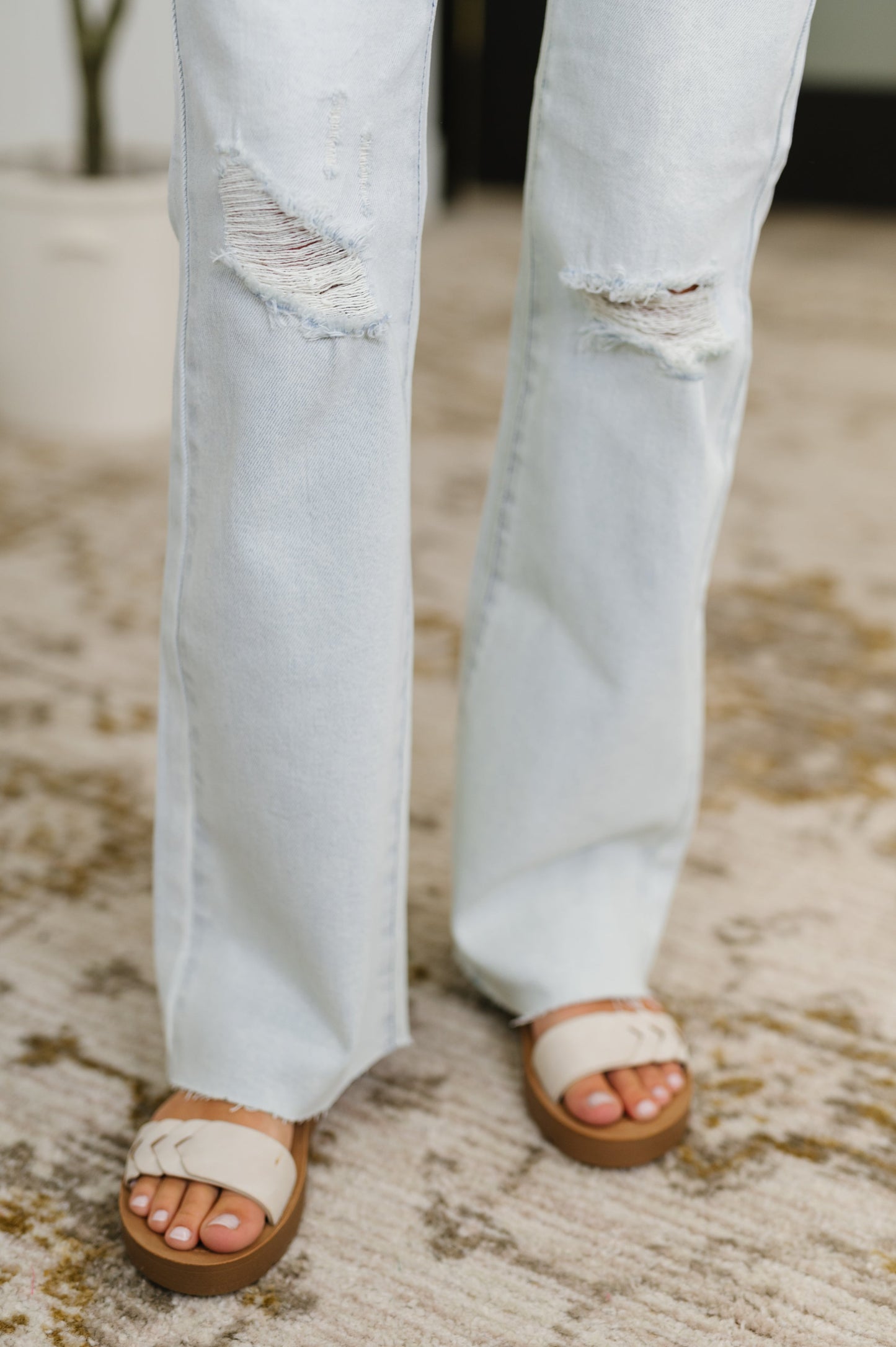 Light blue jeans with distressed details worn by a person, paired with brown sandals on a textured surface.