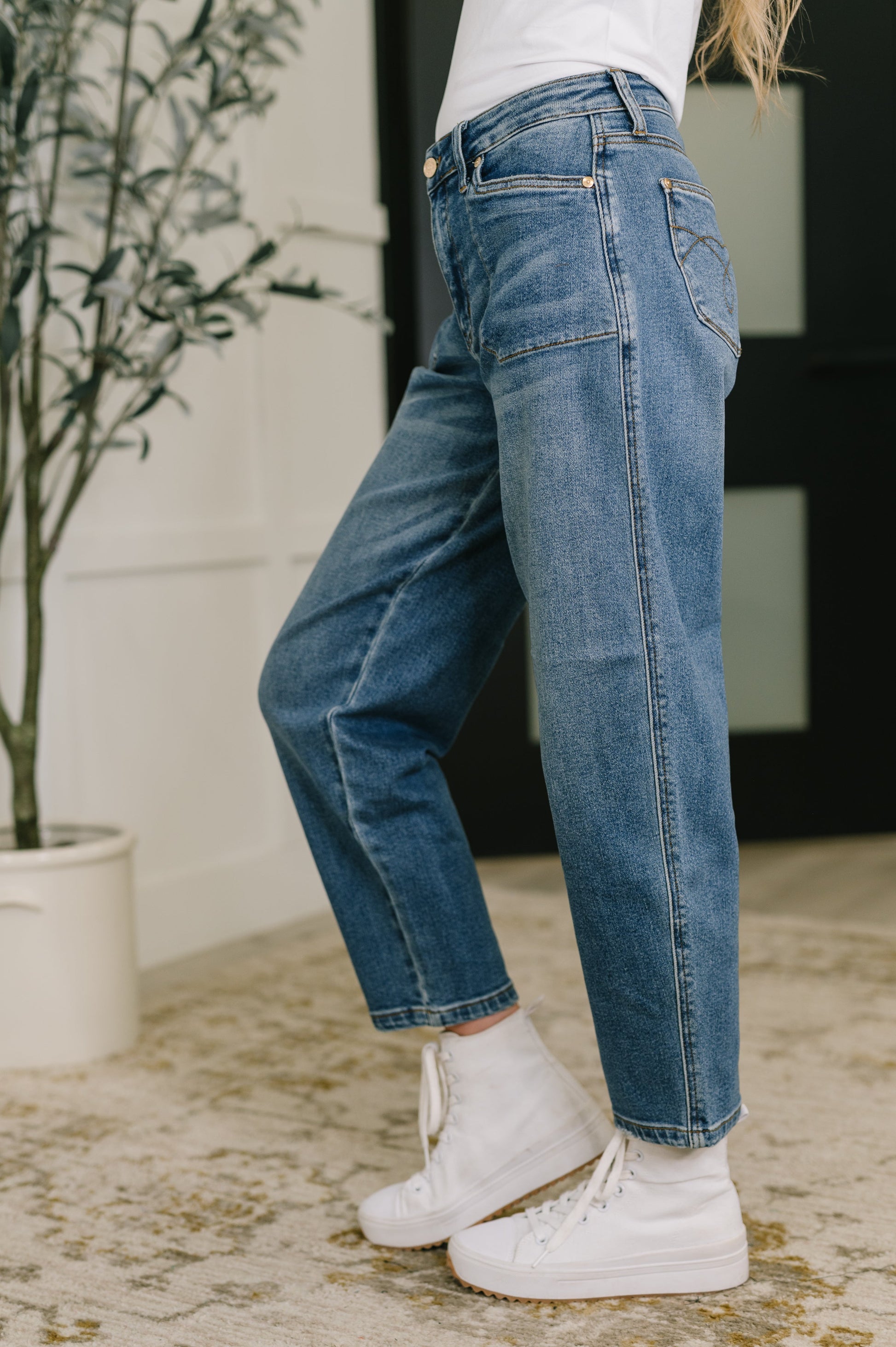 Person wearing blue jeans and white sneakers standing next to a plant indoors.