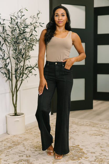 Woman wearing a beige tank top and black flared jeans standing indoors with a plant and wall decor in the background.