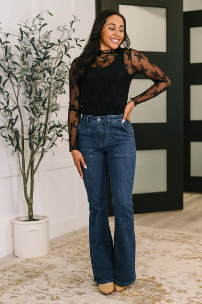 Woman wearing a black sheer top and blue jeans standing indoors with a plant in the background