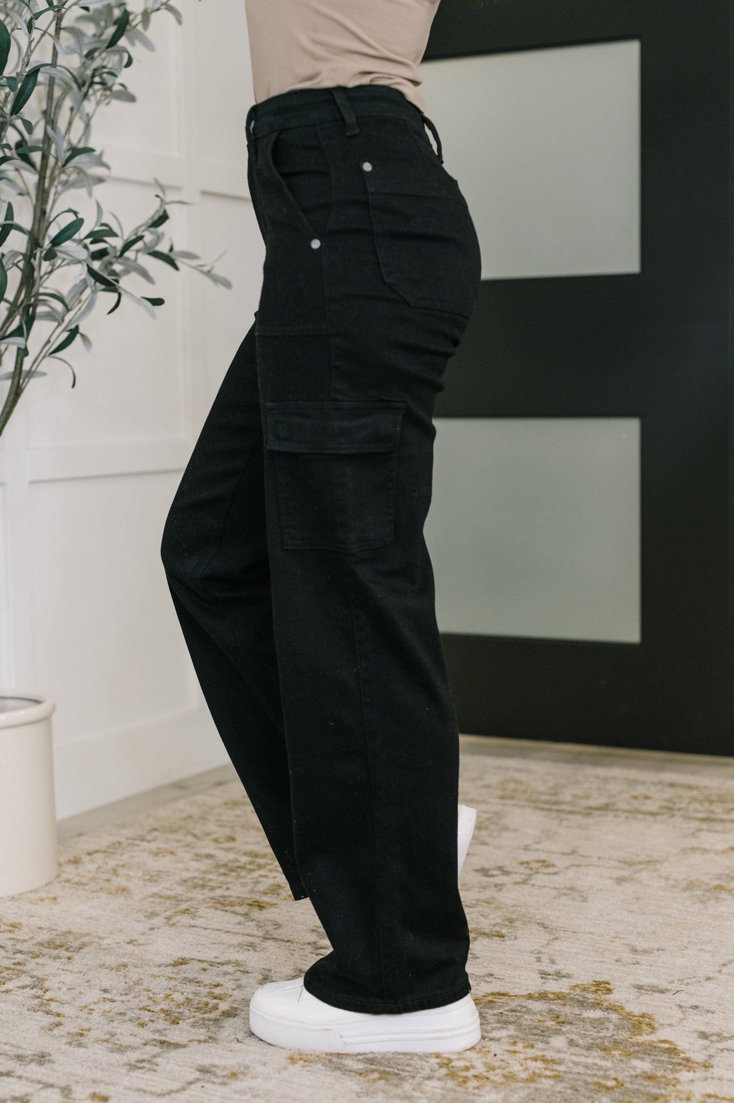 Person wearing black cargo pants standing indoors with a plant and abstract art in the background.